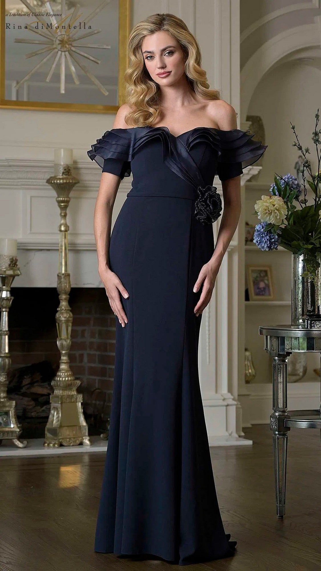 A woman wearing the navy Long Formal Dress: Rina Di Montella RD3418, featuring a fitted sheath silhouette and off-the-shoulder neckline, stands indoors by a fireplace and floral arrangement, looking at the camera.